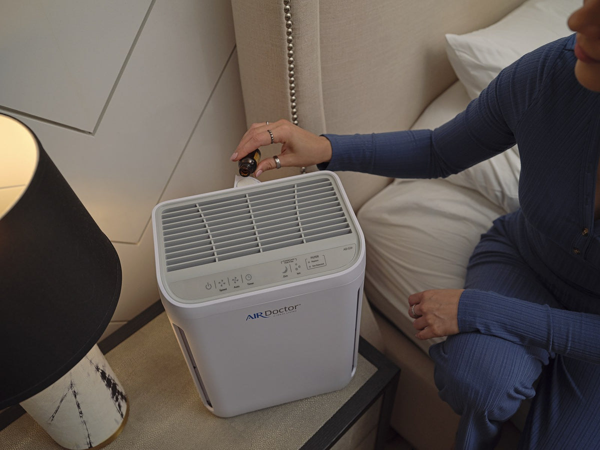 A white AirDoctor portable air purifier sits on a bedside table next to a bed, with a woman in blue pajamas holding a remote control.