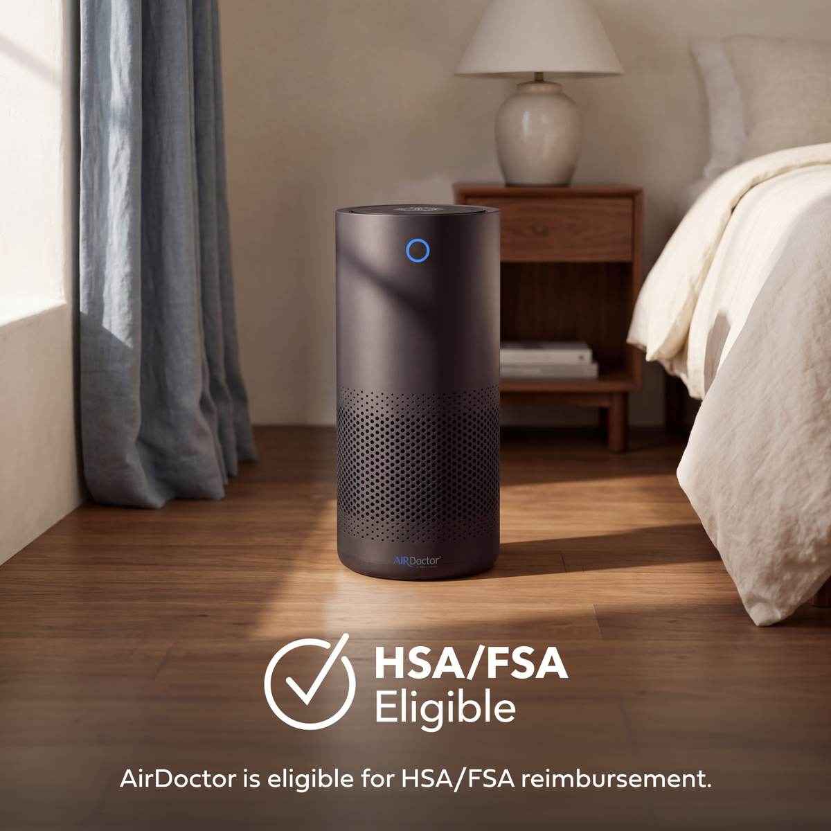 A dark gray cylindrical AirDoctor air purifier stands on a wooden floor in a softly lit bedroom. A blue indicator light glows on top, and the room includes a bed, nightstand, lamp, and curtains.