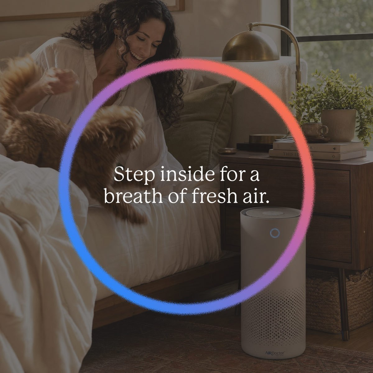 A white portable air purifier with a blue circular indicator light stands on the floor next to a bed in a cozy bedroom setting. A woman is sitting on the bed with a dog, and a wooden nightstand with books and a lamp is visible in the background.