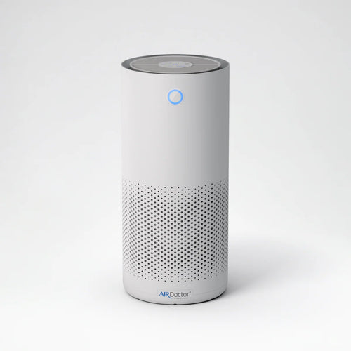A white cylindrical smart air quality monitor with a perforated lower grille, a blue indicator light, and the AIRDoctor logo near the base, set against a clean white background.