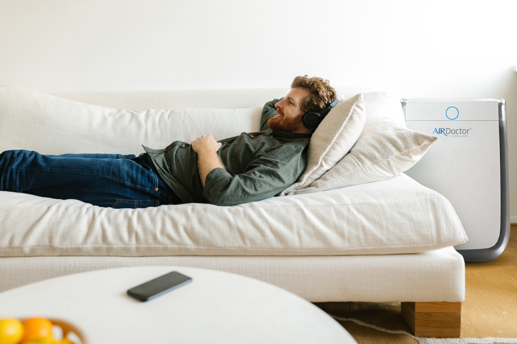A man wearing over-ear headphones lies on a white sofa in a bright living room, with an AIR Doctor air purifier visible beside him.